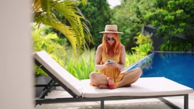Akıllı telefonu kontrol eden bir bayanla tatil sahnesi, yaz tatilinin tadını çıkarmak. Hasır şapkalı kızıl kadın havuz kenarında güneşlenir, meyveli kokteyl yudumlar. Teknoloji ve açık hava rahatlaması arasındaki denge kavramı.