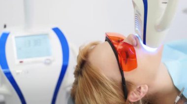 Modern diş muayenehanesinde lazer diş beyazlatan kadın hasta yüzünün yakın çekimi. Koruyucu kırmızı gözlüklü, dişlerini beyazlatan sarışın kadın. Ultraviyole lamba ile diş beyazlatma prosedürü.