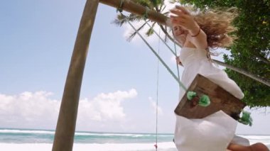 Dalgalı deniz manzarası, eğlenceli tropikal kaçış. Beyaz elbiseli neşeli kadın salıncakta sallanıyor, dalgalı dalgalı saçlar okyanus esintisinde esiyor. Yalnız gezgin, kaygısız anın tadını çıkarır, deniz kenarında sallanır..