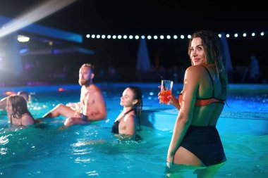 Bikinili güzel bir kadın kokteyl içiyor ve gece havuzu partisinde arkadaşlarıyla takılıyor. İnsanlar lüks bir yüzme havuzunda içkilerle dans edip su sıçratarak eğleniyorlar..