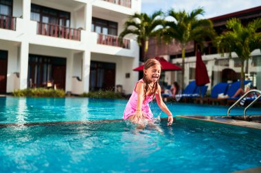 Çocuk yaz tatilinde yüzmeyi, neşeli bir su sıçrayışını seviyor. Pembe mayo giymiş neşeli genç kız tatil köyü havuzuna atladı. Çocuklar aktif eğlence, açık hava aile eğlencesi. Güneşli tatil köşkü. Sıçra, gülümse..