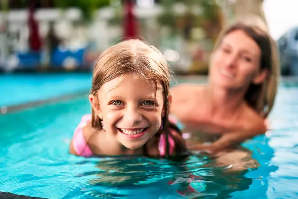 Child first swim lesson under parent supervision. Smiling young girl with swim cap learns swimming in pool, guided by mother. Family, teaching water safety, joyful moments together at summer resort.