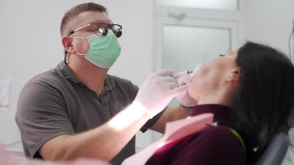 在现代牙科诊所 双筒望远镜的男牙医治疗女病人的牙齿 医生用注射器在牙齿上涂上蓝色蚀刻胶 然后再安装注射器 安装牙冠的过程 — 图库视频影像