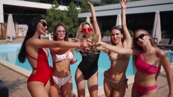 Group of female models pouring and drinking champagne in bright and colorful bikinis at summer party on weekends. Dances at the swimming pool with friends, alcohol, fun and music on hot sunny day.