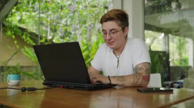 Mutlu bir birey, kolları dövmeli, uzaktan çalışıyor. Modern ofiste dizüstü bilgisayar kullanırken transgender pro gülümser. Kapalı alanda doğal ışık, yeşil bitkiler. Pozitif kapsayıcı iş ortamı. Yavaş çekim.