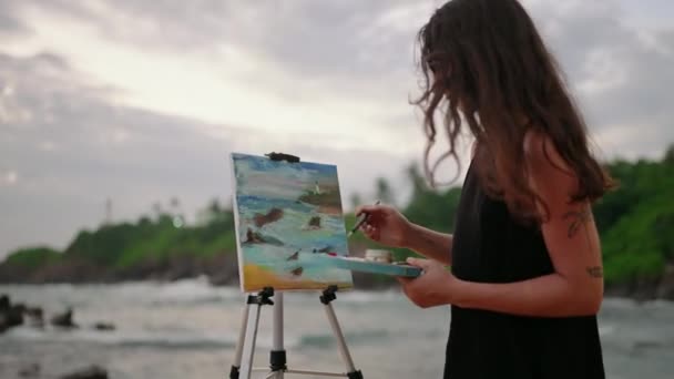 岩の海岸で波が衝突し クリエイティブな女性が海で屋外で働きます アーティストの女性は 夕暮れのキャンバスに嵐のような海の風景を描いています アート インスピレーション 自然はトワイライトビーチの設定でブレンドします スローモーション — ストック動画