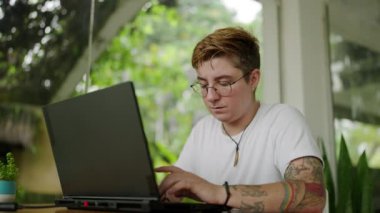 Yeşilliklerle çevrili konsantre profesyonel savunma görevleri. Transseksüel serbest çalışan biri kafede dizüstü bilgisayarda çalışıyor. Seçkin modern çalışma alanı, dövmeli bireyler ekrana odaklı. Yavaş çekim.