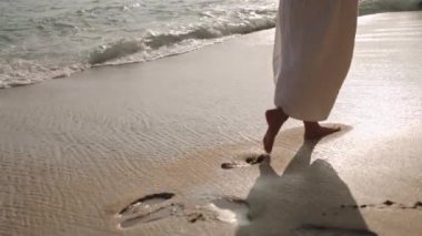 Dalgalar yavaşça ayak izlerini siler. Dalgalı elbiseli çıplak ayaklı insan kumlu sahil şeridinde yürüyor. Gün batımında huzurlu bir sahil yürüyüşü huzurlu bir an ve yansıma sağlar. Yavaş çekim.