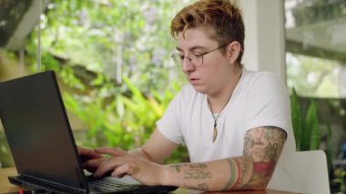 Gözlüklü bireysel odaklanma, kapsayıcılığı ve modern serbest çalışmayı sembolize ediyor. Bitkilerle dolu çalışma alanında dizüstü bilgisayarda dövmeli transseksüel profesyonel tipler. Yavaş çekim.