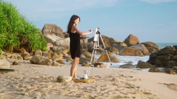 波の衝突 熱帯の風が吹く一方 熟練した女性は芸術を通して沿岸の風景の本質を捉えます クリエイティブな女性アーティストは キャンバスに海の景色を描く砂浜に裸足で立っています スローモ — ストック動画