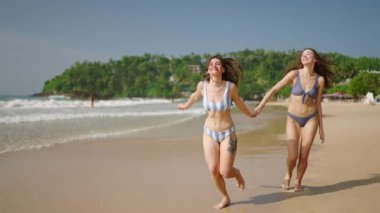 Bikinili mutlu kızlar el ele tutuşup tropikal adada kumsalda koşuyorlar. Gülümseyen beyaz kadınlar okyanus kıyısında gezi turunda eğleniyorlar. Mayolu bayan arkadaşlar yaz tatilinin tadını çıkarın..