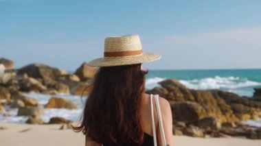 Tek başına gezen kumlu sahilde gezer. Hasır şapkalı kadın resimli deniz günbatımının tadını çıkarıyor. Dalgalar kayalara çarpar. Kadın okyanus ufkuna bakıyor, altın saat ışığında gülümsüyor. Sakin sahil yürüyüşü.