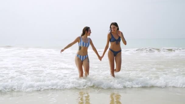 快乐的女孩手牵着手 走出海水 穿着比基尼的苗条年轻貌美的女人在海里洗过澡后就会出水 女性朋友笑着 在暑假散步 慢动作 — 图库视频影像