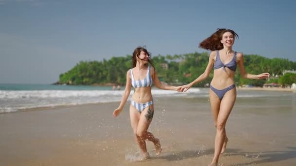 年轻女子穿着泳衣 手牵着手 沿着海水边奔跑 微笑着的漂亮姑娘们跑着 让水在海滨飞溅 两名女游客在天堂岛上的海滨玩得很开心 — 图库视频影像