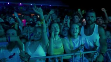 2021-08-06 - Mariupol Şehir Festivali, Ukrayna. Hayranlar konserden hoşlanır, neon ışıklar yüzleri aydınlatır. Gece müzik festivalinde enerjik kalabalık dans ediyor. Arkadaşlar selfie çekerler, sahne performansından heyecan duyarlar..