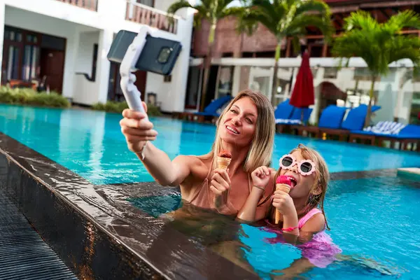 Mom uses selfie stick for live stream video. Mother and daughter enjoy ice cream while swimming in resort pool. Happy family vacation moment with tech interaction. Mom blogs, child smiles with treat.