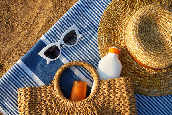 Woven bag with sunscreen inside next to stylish sun hat. Summer accessories on sandy beach at sunset. White sunglasses lie on blue striped towel. Vacation essentials for sun protection.