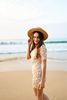 Arka planda mavi okyanus dalgaları. Vintage çiçekli elbise ve hasır şapkalı gülümseyen kadın kumlu sahilde yürüyor. Yaz, moda, eğlence konsepti. Mutlu kadın deniz kenarında günbatımını sever, eski usul..