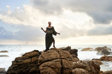 İkili olmayan siyah tanrısal kişi okyanustaki kayaların üzerinde zarifçe duruyor. Rock plajında lüks bir elbise ve mücevherle gezen trans etnik model. Lgbtq. Gurur ayı