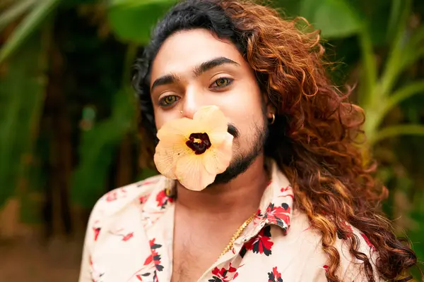 Hawaiian shirt blends with green background, representing LGBT pride, ethnic diversity. Dark-skinned man with curly long hair poses in nature, flower in mouth expressing confidence, non-conformity.