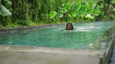 Yeşil yapraklarla çevrili suda dinlenirler. Çok ırklı bir kadın orman havuzunda yüzüyor. Boş zaman aktivitesi, yaz günü ferahlatıcı. Sakin bir yüzüş, doğal güzelliğin tadını çıkarıyor. Yavaş çekim.