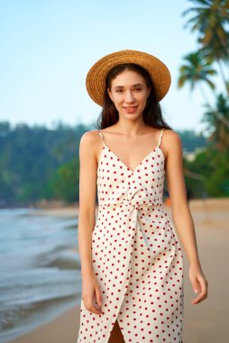 Retro yaz modası sergileniyor. Puantiyeli elbise ve hasır şapkalı kadın güneşli sahilde gülümsüyor. Dişi deniz kenarından, mavi gökyüzü ve yumuşak dalgalarla tropikal arkaplanlardan hoşlanır. Tatil sahnesi, rahat bir hava..