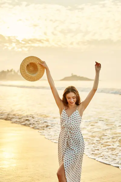 Arms raised, holding wide-brimmed hat, she smiles, embodying leisure, travel, and seaside freedom. Elegant woman in polka dot dress enjoys sunset at beach, waves gently wash over her bare feet.