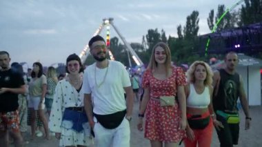 2021-08-07 - Mariupol Şehir Festivali, Ukrayna. Moda kıyafetleri içinde bir grup, güneş gözlükleri etkinliğin tadını çıkarıyor, dönme dolabın ışıkları arka planda. Moda arkadaşları güler, müzik festivalinde yürürler, akşam.