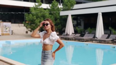 Lüks bir kulüpte etek, bluz ve güneş gözlükleriyle yüzme havuzunda poz veren ve gülümseyen güzel bir kız modeli. Yaz partisinde çekici bir kadın göletin berrak mavi sularının yanında tatil yapıyor..
