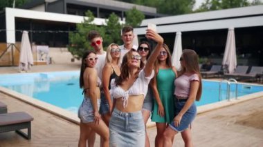 Bir grup kadın ve erkek arkadaş lüks plaj klübü restoranında yüzme havuzunda telefonda selfie çekiyorlar. Kızlar ve erkekler yaz kıyafetleri içinde pov videosu için poz veriyor ve gülümsüyorlar..