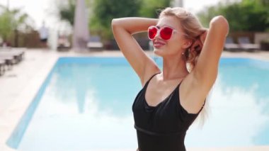 Sarı saçlı kadın kırmızı güneş gözlüğü, şort ve yüzme havuzunda güneş pansiyonları ve çam ağaçlarıyla fotoğraf çekmek için dans ediyor. Tatillerde poz vermek ve flört etmek.