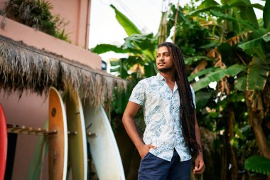 BIPOC coach awaits students for lesson by lush green plants. Confident dark-skinned surf instructor stands among colorful boards in tropical setting. Surf camp mentor ready for day training sessions. clipart