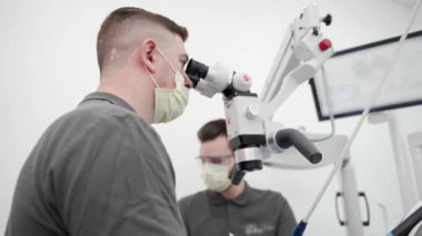 Modern diş kliniğinde mikroskop kullanarak hasta dişini ameliyat eden bir erkek diş hekimi. Diş hekimi diş kanalını temizliyor ve diş sinirlerini dağlıyor. Doktor hastalara kanal tedavisi uyguluyor..