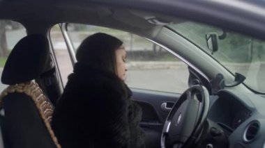 Kadın sürücü şehir turu için hazırlanıyor, dikiz aynası ayarlanıyor, ateşleme hazır. Paltolu kadın arabaya biniyor, işe gitmeden önce emniyet kemerini takıyor. Otomobil yolculuğu, kişisel ulaşım, sürücü güvenliği.