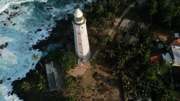 象徴的なビーコンの近くの岩の海岸に波が衝突する ドローンの歴史的なドローン灯台 活気に満ちた熱帯の葉 白い砂浜 澄んだ青い海水 — ストック動画