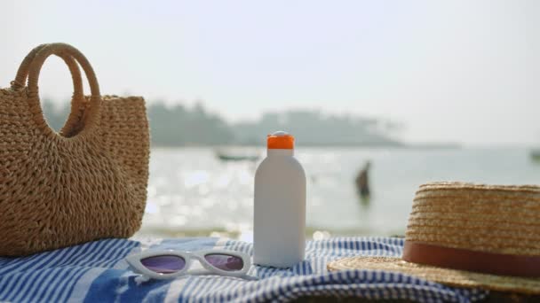 皮膚保護のための日焼け止めのボトル 晴れた日には海の近くのストライプタオルに置かれたビーチの必需品 カジュアルな夏のアクセサリーには ウィッカーバッグ サングラスが含まれています — ストック動画