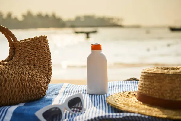 Bottle of SPF lotion ready for skin protection. Beach scene with sun cream, hat, sunglasses, and straw bag on towel. Summer accessories lie on sandy shore, with ocean waves in background.