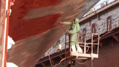 Sprey tabanca kırmızı kaplama sürerken koruyucu giysi. İşçi tersanede gövde boyuyor, büyük bir geminin bakımını yapıyor. Denizcilik endüstrisi ve gemi yenileme süreci iş başında sergilendi.