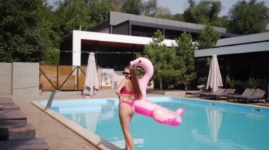 Pembe bikinili, güneş gözlüklü, yüzme havuzunda poz veren, şişme flamingo tüplü güzel bir manken. Uzun saçlı kadın tatil için şehir plaj kulübünde dinleniyor..