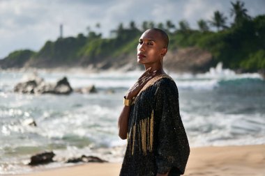 Lgbtq siyah model lüks elbiseler içinde karamsar görünümlü, okyanus manzaralı sahilde poz veriyor. Uzun gösterişli elbiseler ve aksesuarlar içinde ikili olmayan etnik model kameraya küstahça bakar, boynuna dokunur..