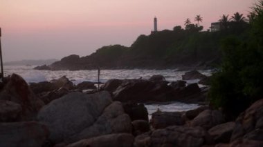 Dalgalar alacakaranlıkta kayalık kıyıya çarpar. Deniz feneri, pamuk helvanın pembe günbatımına karşı tepede duruyor. Sahil manzarası navigasyonu yansıtır, deniz güzelliğini, sakin akşam renkleriyle. Yavaş çekim.