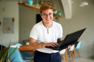 Inclusive workplace, young trans person enjoys career, experiences job fulfillment with tech in eco-friendly area. Smiling transgender professional holds laptop, works happily in modern office space. clipart