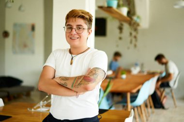 Dövmeli kollar çapraz, iş yerinde mutlu ve arka planda yoğun bir ekip var. Transseksüel erkek profesyonel gülümsemesi, açık ofis alanında duruyor. LGBTQ bireyleri için pozitif iş yeri.