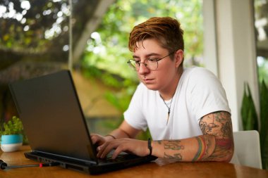 Uzaktan kumandalı dövme teknolojisi olan odaklanmış bir birey. Transseksüel serbest çalışan biri, tropikal çalışma alanında dizüstü bilgisayarla çalışır. Açık, kapsayıcı iş tesisleri. Yaratıcılık, verimlilik, LGBTQ dostu ortam.