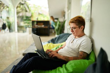 Rahat bir ortam, ışık dolu bir oda, modern iş hayatı dengesi, işyerine dahil edilme. Laptopta transseksüel profesyonel çalışmalar, yeşil fasulye torbası üzerine oturmuş, gündelik ofis alanı..