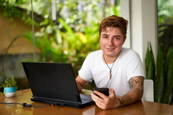 stock image Smiling, pro, connected, inclusive work environment. Transgender individual works remotely, multitasking with laptop, smartphone at home office. Casual biz, wireless tech, body art visible.