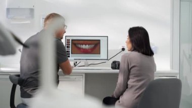 Kozmetik dişçisi modern diş kliniğindeki kadın hastaya gülümseme fotoğrafı gösteriyor. Stomatolog ve hasta kaplama modellemesini tartışıyor. Doktor kadınlara diş fotoğraflarını açıklıyor..