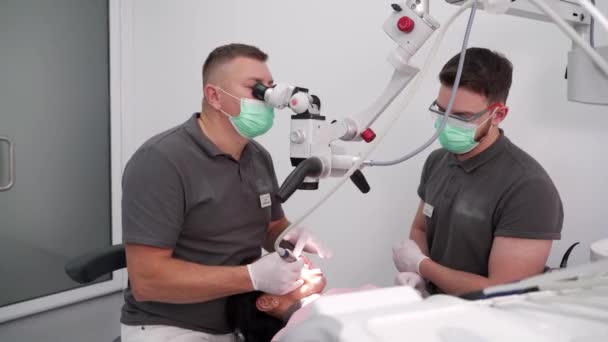 男性歯科医は 現代の歯科オフィスで患者の歯を治療するために顕微鏡を使用してアシスタントと協力しています 医者は顕微鏡を使用し ベニヤを取付けるために歯のエナメルを取除きます ストロマティスト トリートメント キャリー — ストック動画