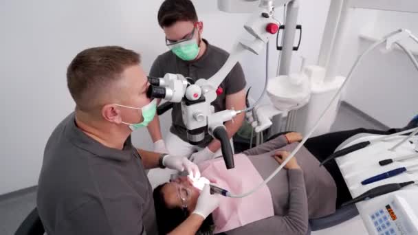 男性歯科医は 現代の歯科オフィスで患者の歯を治療するために顕微鏡を使用してアシスタントと協力しています 医者は顕微鏡で働き ベニヤを取付けるための歯のエナメルを取除きます コスメティック歯科 — ストック動画