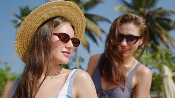 幸せな若い女性は友人が熱帯のビーチでサンクリームを塗るのを助けます 陽気な女の子は 楽園の島 ヤシの木で日焼けする 女性の友人がローションを使用しています クローズアップ — ストック動画